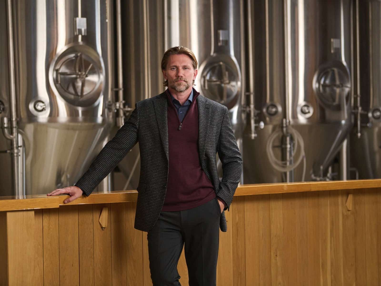 A man in a grey blazer and burgundy sweater leaning on a bar in a brewery
