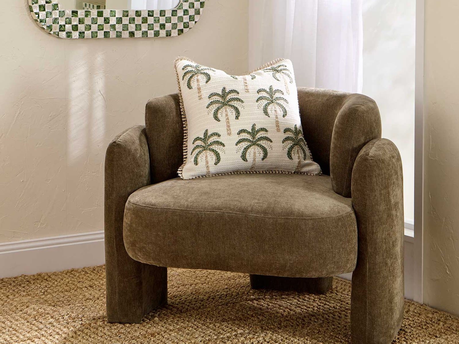 A rounded olive green upholstered occasional chair with a palm-embroidered cushion on a jute rug, beside a checked-frame mirror and sheer curtains.