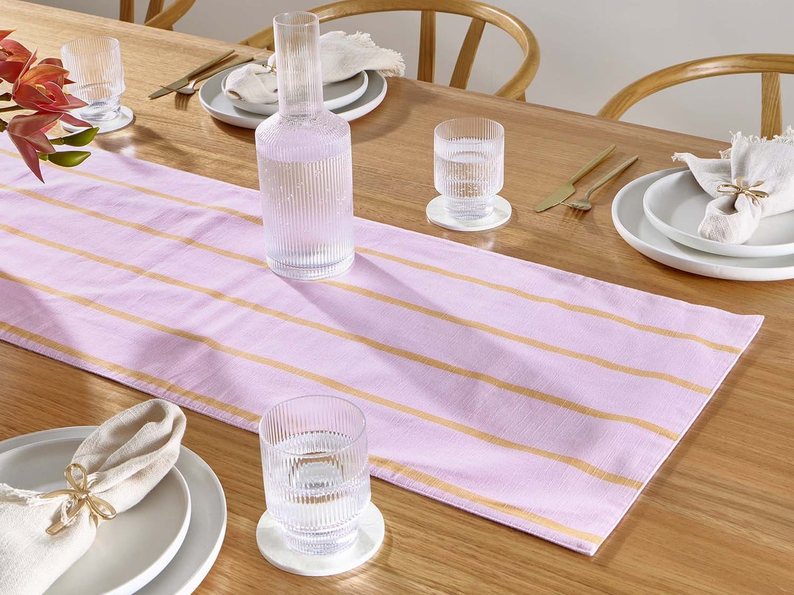 A pink and gold striped reversible table runner down a timber dining table set with grey plates, linen napkins, ribbed glasses and a glass carafe.