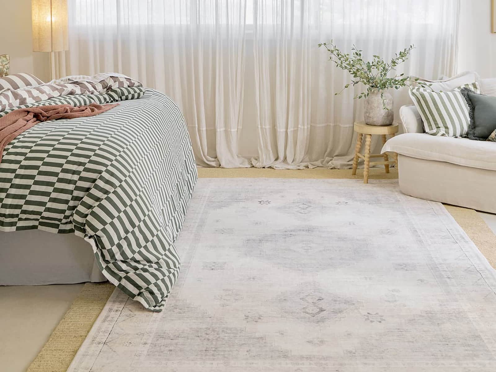 A pale distressed-pattern washable floor rug in a bright bedroom beside a bed with a green and white checked quilt and a white sofa by sheer curtains.