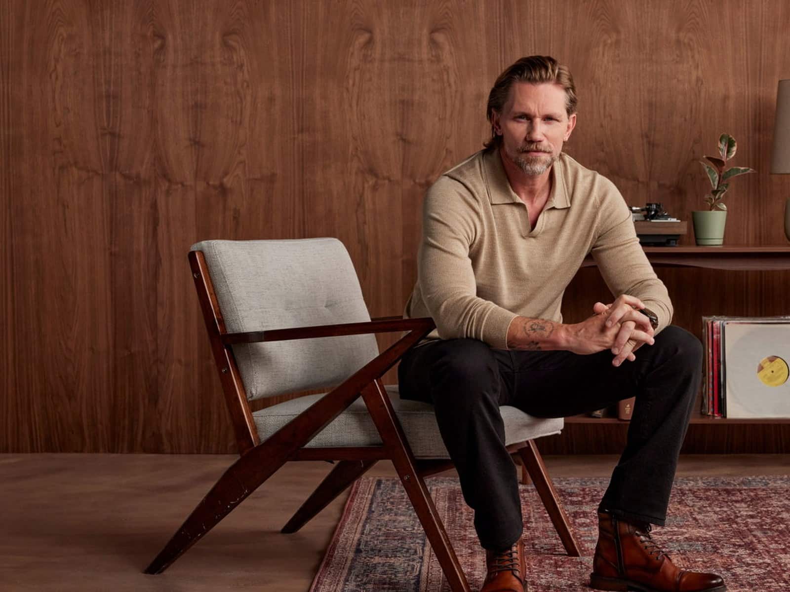 A man in a cream knit polo and dark trousers seated in a mid-century armchair