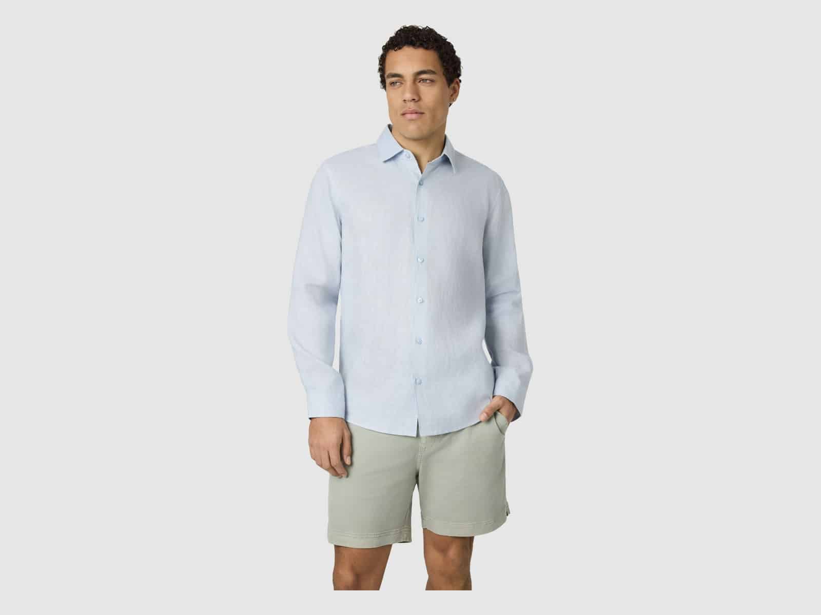A man wearing the Connor Hamilton Linen Shirt in Light Blue with sage shorts, one hand in his pocket