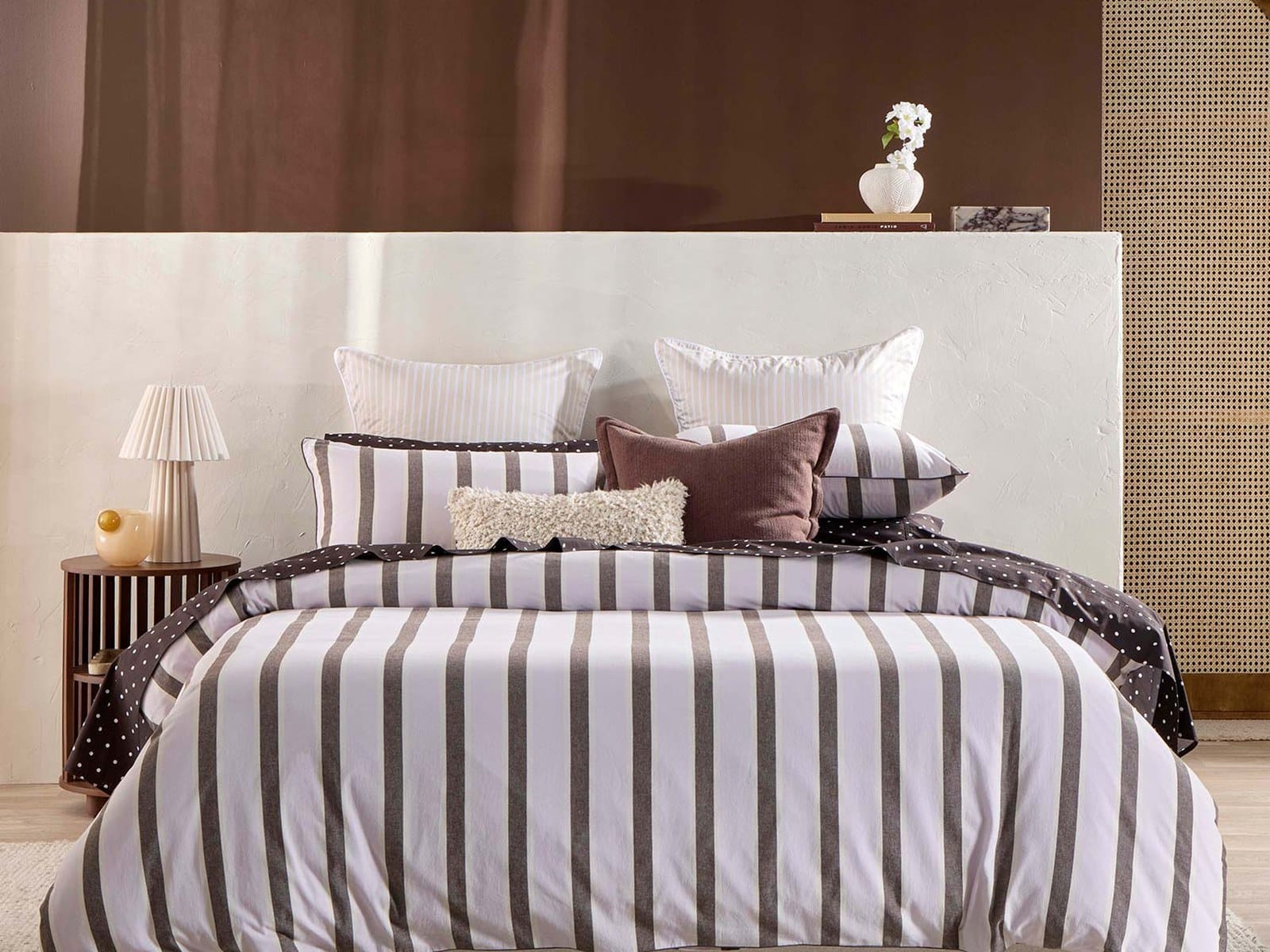 A made bed with a chocolate and lilac striped Pillow Talk quilt cover, polka dot sheets, striped pillowcases and a brown linen cushion, against a cream headboard wall with a pleated lamp on a timber side table.